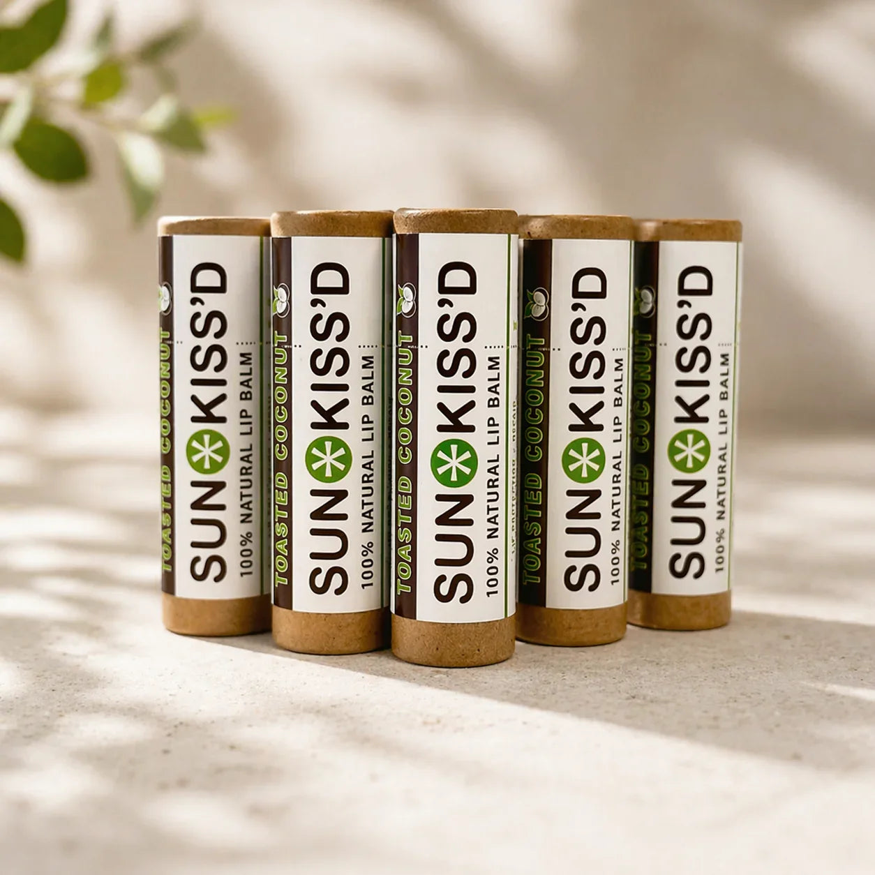 Five Sunkiss'd toasted coconut natural lip balm tubes on a beige surface with soft lighting.