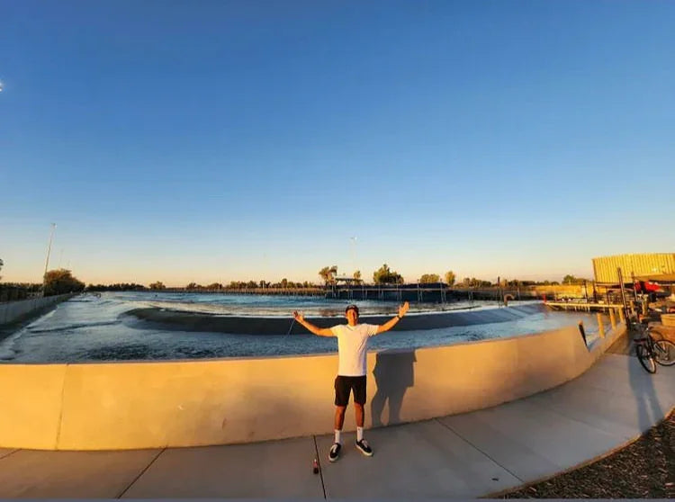 Person standing with arms raised in front of a large outdoor wave pool at sunset